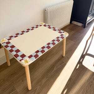 May include: A rectangular children's table with a cream-colored top and light wood legs. The table's border features a checkered pattern in burgundy and light blue, with yellow star accents at the corners. The table is approximately 30 inches long.