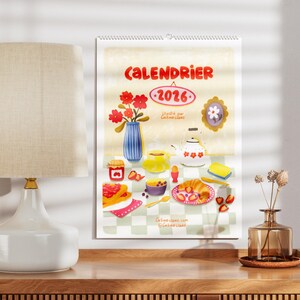 Op de afbeelding: Een kalender voor 2026 met een grillige illustratie van een ontbijt, met een vaas met rode bloemen, een theepot en diverse etenswaren. Het woord "CALENDRIER" staat bovenaan.