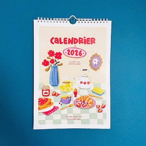 Op de afbeelding: Een 2026-kalender met spiraalbinding, met een kleurrijke illustratie van een ontbijt. De afbeelding bevat een vaas met rode bloemen, een theepot en een bord met eten. Tekst op de kalender luidt "CALENDRIER 2026" en "Callmeciano".