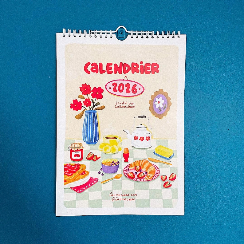 Op de afbeelding: Een 2026-kalender met spiraalbinding, met een kleurrijke illustratie van een ontbijt. De afbeelding bevat een vaas met rode bloemen, een theepot en een bord met eten. Tekst op de kalender luidt "CALENDRIER 2026" en "Callmeciano".