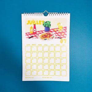 Op de afbeelding: Een spiraalgebonden kalender met een juli-thema. De illustratie toont een picknicksc&egrave;ne met eten, drinken en het woord "JUILLET". Het kalenderraster is geel met data. De achtergrond is effen blauw.