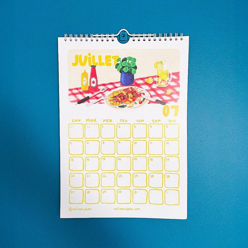 Op de afbeelding: Een spiraalgebonden kalender met een juli-thema. De illustratie toont een picknicksc&egrave;ne met eten, drinken en het woord "JUILLET". Het kalenderraster is geel met data. De achtergrond is effen blauw.