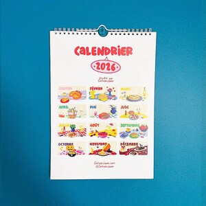 Op de afbeelding: Een kalender voor 2026 met een witte achtergrond en een blauwe metalen clip. De kalender bevat kleurrijke illustraties voor elke maand, met het woord "CALENDRIER" in het rood bovenaan. De naam en website van de kunstenaar staan onderaan.