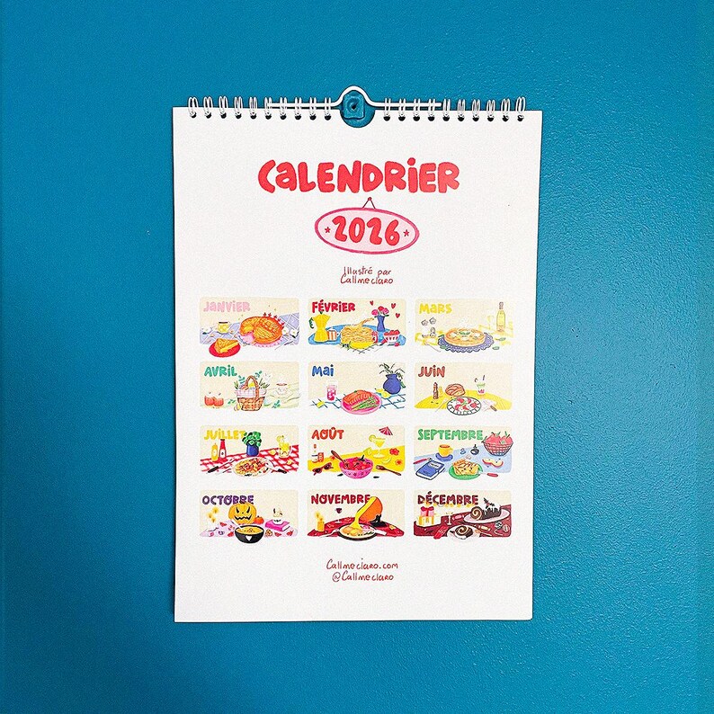 Op de afbeelding: Een kalender voor 2026 met een witte achtergrond en een blauwe metalen clip. De kalender bevat kleurrijke illustraties voor elke maand, met het woord "CALENDRIER" in het rood bovenaan. De naam en website van de kunstenaar staan onderaan.