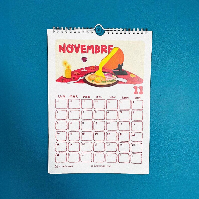 Op de afbeelding: Een spiraalgebonden kalender voor november met een kleurrijke illustratie van een fondue-sc&egrave;ne. De kalender heeft een rasterindeling met data en dagen van de week in het Frans. De tekst "NOVEMBRE" staat bovenaan, met het nummer "11" in rood.