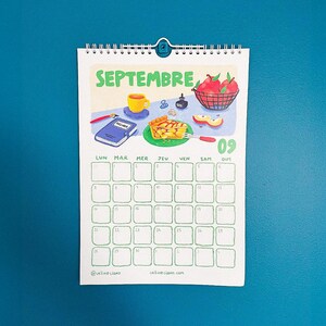 Op de afbeelding: Een kalender voor september met illustraties van taart, appels en een kopje koffie. De kalender heeft een blauwe omslag en een teal achtergrond. De dagen van de week staan in het Frans. Spiraalgebonden.