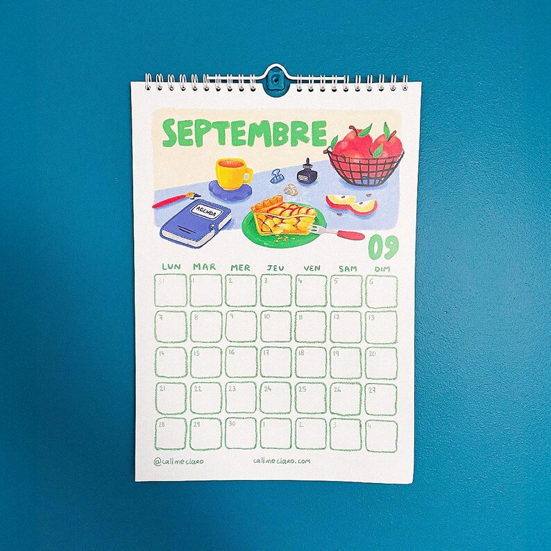 Op de afbeelding: Een kalender voor september met illustraties van taart, appels en een kopje koffie. De kalender heeft een blauwe omslag en een teal achtergrond. De dagen van de week staan in het Frans. Spiraalgebonden.