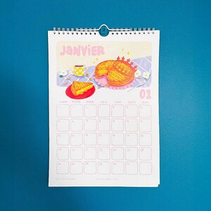 Op de afbeelding: Een spiraalgebonden kalender voor januari, met een kleurrijke illustratie van een taart en een kopje koffie. Het woord "JANVIER" staat bovenaan, met de datum "01" in roze. De achtergrond is blauw.