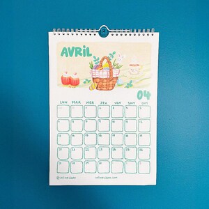 Op de afbeelding: Een spiraalgebonden kalender voor april, met een aquarel illustratie van een paasmand, eieren en een kopje thee. Het woord "AVRIL" staat bovenaan, met de dagen van de week en de data eronder. De kalender staat tegen een tealblauwe achtergrond.