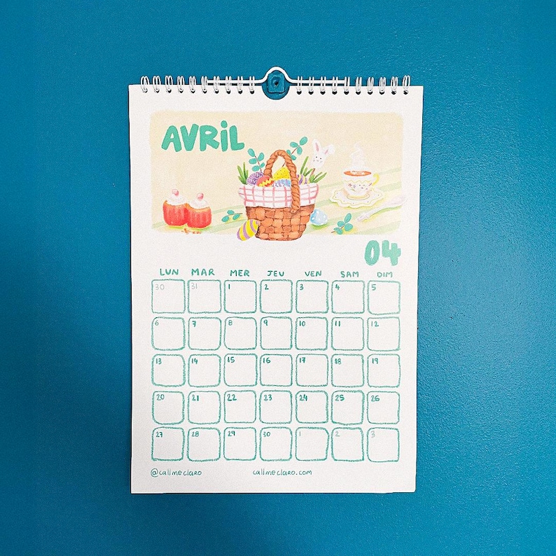 Op de afbeelding: Een spiraalgebonden kalender voor april, met een aquarel illustratie van een paasmand, eieren en een kopje thee. Het woord "AVRIL" staat bovenaan, met de dagen van de week en de data eronder. De kalender staat tegen een tealblauwe achtergrond.
