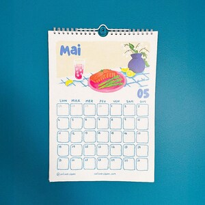 Op de afbeelding: Een spiraalgebonden kalender voor de maand mei, met een grillige illustratie van een maaltijd met een drankje, zalm en asperges. De kalender heeft een blauw en wit raster met data en dagen van de week. De tekst "Mai" staat bovenaan.