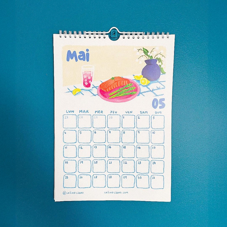 Op de afbeelding: Een spiraalgebonden kalender voor de maand mei, met een grillige illustratie van een maaltijd met een drankje, zalm en asperges. De kalender heeft een blauw en wit raster met data en dagen van de week. De tekst "Mai" staat bovenaan.