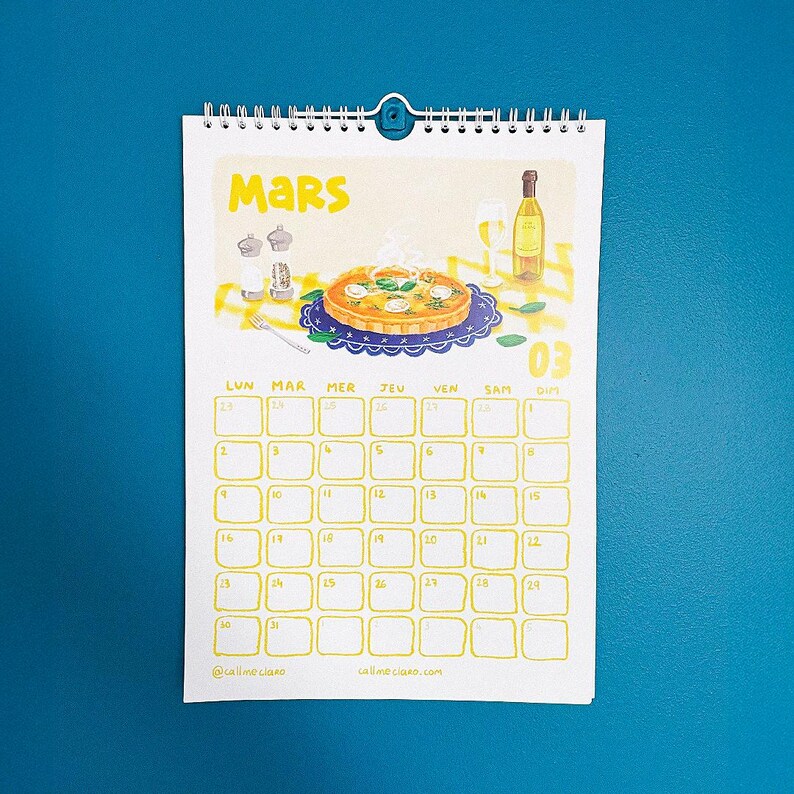 Op de afbeelding: Een spiraalgebonden kalender voor maart, met een gele illustratie van quiche, wijn en een glas. De kalender heeft een blauwe achtergrond en een raster met gele data. De tekst "Mars" staat bovenaan.