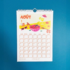 Op de afbeelding: Een spiraalgebonden kalender met de maand augustus, ge&iuml;llustreerd met een fruitschaal, zonnebril en een cocktail. De kalendergrid is lichtbruin, met de dagen van de week en datums zichtbaar. De tekst "Ao&ucirc;t" staat bovenaan.