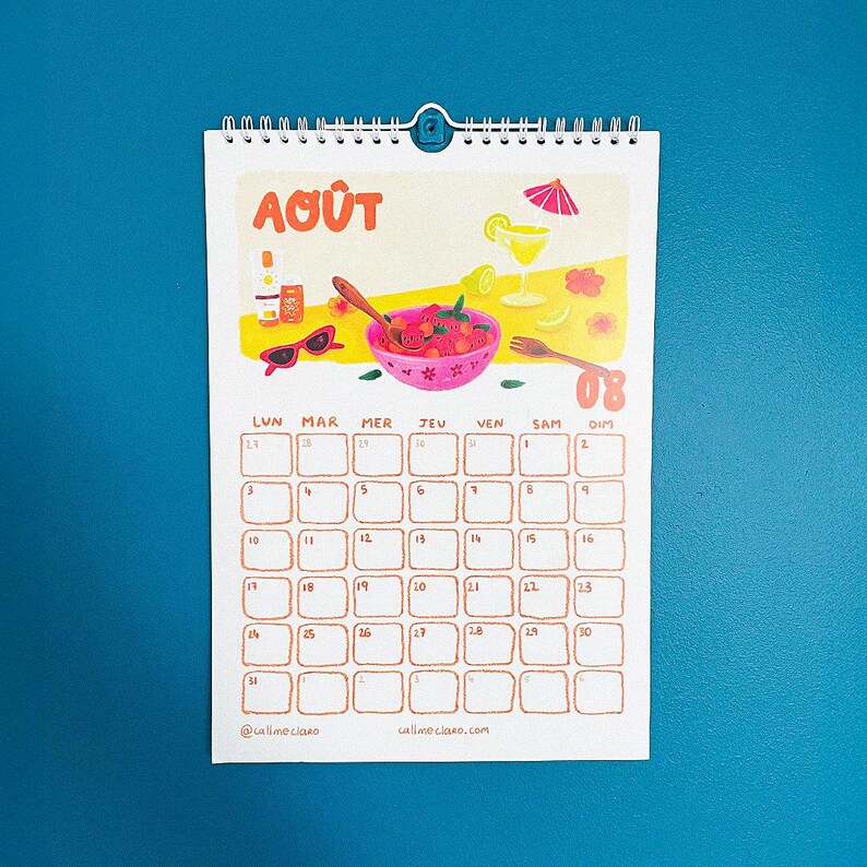 Op de afbeelding: Een spiraalgebonden kalender met de maand augustus, ge&iuml;llustreerd met een fruitschaal, zonnebril en een cocktail. De kalendergrid is lichtbruin, met de dagen van de week en datums zichtbaar. De tekst "Ao&ucirc;t" staat bovenaan.