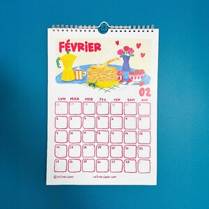 Op de afbeelding: Een spiraalgebonden kalender voor februari met een kleurrijke illustratie van ontbijt. Het woord "F&Eacute;VRIER" staat bovenaan. De kalendergrid is rood, met de dagen van de week in het Frans. Onderaan staat "@callmeclaro" en "callmeclaro.com".