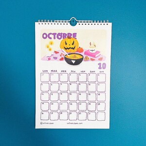Op de afbeelding: Een spiraalgebonden wandkalender voor oktober, met een Halloween-thema. De kalender heeft de paarse titel "OCTOBRE" en illustraties van een pompoen, kaarsen en een kom soep. De kalendergrid is paars.