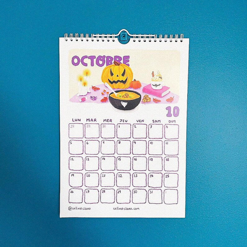 Op de afbeelding: Een spiraalgebonden wandkalender voor oktober, met een Halloween-thema. De kalender heeft de paarse titel "OCTOBRE" en illustraties van een pompoen, kaarsen en een kom soep. De kalendergrid is paars.