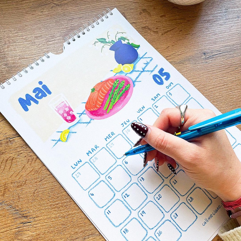 Op de afbeelding: Een spiraalgebonden kalender met de maand "Mai" in blauwe letters. De kalender bevat illustraties van eten en drinken. Een hand met een blauwe pen schrijft op de kalender. De kalender heeft een raster van data.