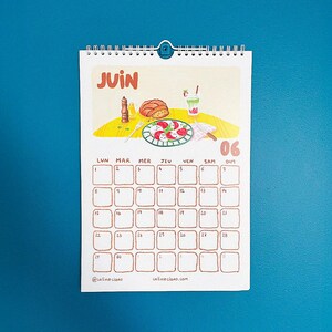 Op de afbeelding: Een spiraalgebonden kalender voor juni met een illustratie met eten als thema. De kalendergrid is bruin, met de dagen van de week in het Frans. De datum 06 is gemarkeerd. De naam en website van de kunstenaar staan onderaan.