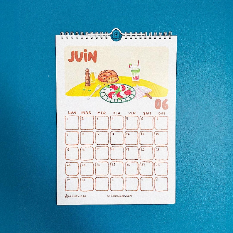 Op de afbeelding: Een spiraalgebonden kalender voor juni met een illustratie met eten als thema. De kalendergrid is bruin, met de dagen van de week in het Frans. De datum 06 is gemarkeerd. De naam en website van de kunstenaar staan onderaan.