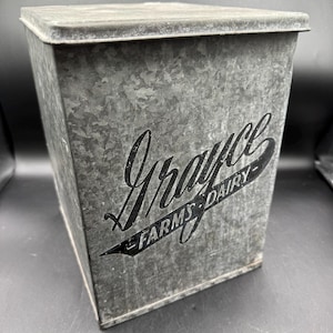 May include: A rectangular galvanized metal container with a lid, featuring a mottled gray finish. The front displays black printed text that reads "Grayce" in a large script font, with "FARMS DAIRY" written in a smaller sans-serif font below.