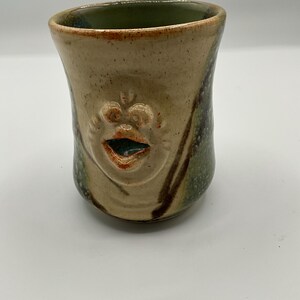 May include: A handmade ceramic mug with a face design. The mug is tan with green and brown accents. The face has a wide open mouth and is centered on the mug. The inside of the mug is green.