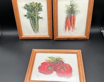 May include: Three framed watercolor prints featuring vegetables. One print depicts celery, another carrots, and the third, red bell peppers. The frames are made of light brown wood, and the artwork is on white paper.