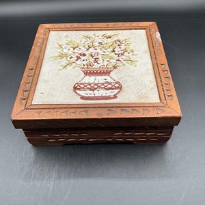 Puede incluir: Caja de madera rectangular con tapa de azulejo floral. El azulejo presenta un jarrón con flores blancas y hojas verdes. La caja es marrón con detalles tallados en los bordes y la base.