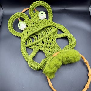 May include: A handmade green macrame frog wall hanging with a bamboo ring base. The frog has large, round eyes and a textured, open weave design. The frog's eyes are white with green details.