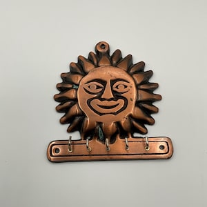 May include: Copper sun-shaped key holder with a smiling face and radiating sun rays. The key holder has four hooks and a small hole at the top for hanging. The key holder is made of copper and has a rustic appearance.