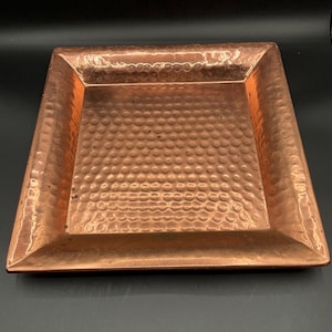 May include: A square copper tray with a hammered texture. The tray has a raised edge and a reddish-brown color. The surface is covered in small, evenly spaced dimples. The tray is likely used for serving or display.