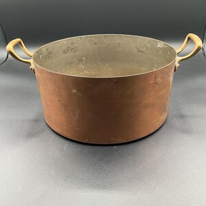 May include: A large copper pot with two brass handles. The pot is empty and has a shiny, slightly tarnished surface.