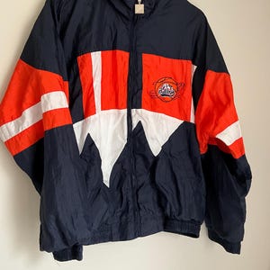 May include: A navy blue and orange vintage jacket with white accents. The jacket features a full zipper, a stand-up collar, and a logo on the chest. The sleeves have white stripes and the jacket is hanging on a black hanger.