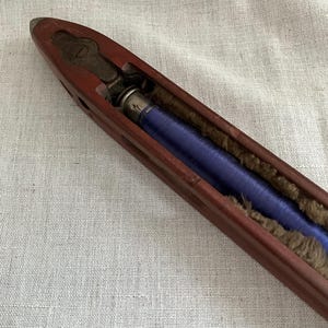 May include: A wooden bobbin with a blue thread wound around it. The bobbin is open on one side, revealing the thread.