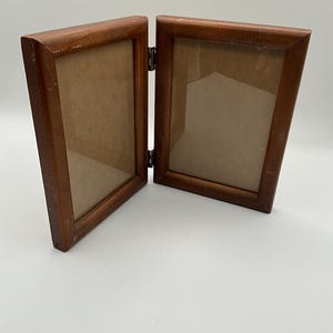 May include: A hinged, wooden double picture frame. The frame is made of dark brown wood and has two rectangular openings with a light brown backing. The frame is open, revealing the hinge that connects the two sides.