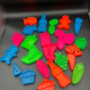 May include: A collection of colorful plastic food-shaped cookie cutters. The cutters include shapes of fish, bottles, carrots, grapes, apples, and cupcakes. The cutters are in shades of blue, green, orange, pink, and red.