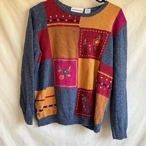 May include: A blue, red, orange and pink patchwork sweater with embroidered flowers. The sweater has a crew neck and long sleeves. The brand label reads "Alfred Dunner".