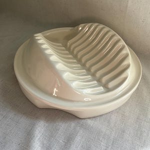 May include: Off-white ceramic egg serving dish. The dish has a rounded base with a ridged, curved section for holding the eggs. The dish is designed to hold hard-boiled eggs and is a kitchenware item.