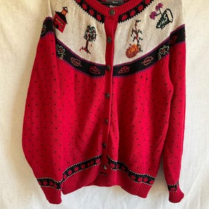 Könnte beinhalten: Roter Cardigan mit Rundhalsausschnitt und Knopfverschluss. Der cremefarbene Einsatz ist mit Stickereien von Haus, Bäumen und Blumen verziert. Der Körper des Pullovers ist rot mit kleinen schwarzen herzförmigen Mustern.