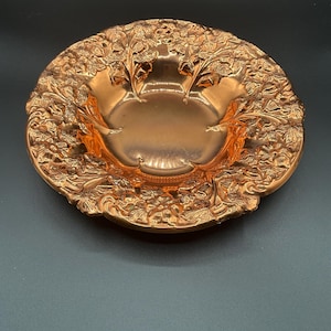 May include: A copper bowl with an ornate floral design. The bowl is round and has a slightly raised rim. The floral design is embossed and covers the entire surface of the bowl.