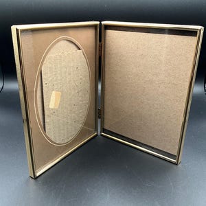 Puede incluir: Un marco de fotos de metal dorado con dos aberturas. El marco está abierto y la parte posterior del marco es visible. La parte posterior del marco está hecha de cartón.