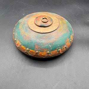 May include: A round, antique wooden box with a lid. The box is painted in shades of green, orange, and brown, with a metal ring on top. The edges are adorned with small, decorative metal studs. The box appears to be handcrafted.