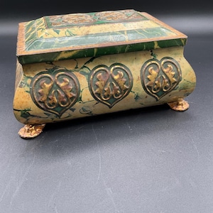 May include: A vintage green and gold tin box with a marbled design and three heart-shaped accents. The box has four feet and a hinged lid.