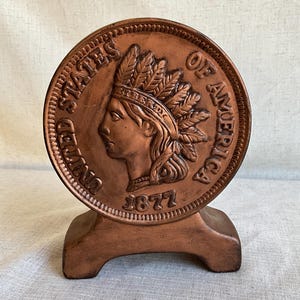 May include: A copper-colored ceramic coin bank shaped like a large coin with the words "United States of America" and the year "1877" on it. The coin features a profile of a Native American woman wearing a headdress with feathers.