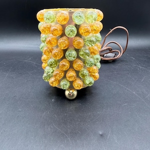 May include: A vintage table lamp with a cylindrical shade. The shade is adorned with a pattern of orange and green glass bubble-like embellishments. The lamp has a gold-colored base and a brown electrical cord.