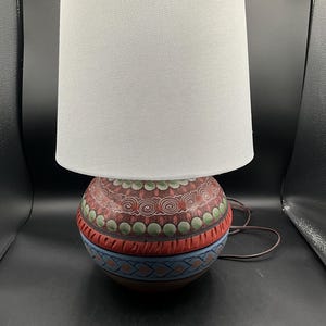 Könnte beinhalten: Dekorative Tischlampe mit weißem Lampenschirm und rundem, handbemaltem Sockel. Der Sockel hat ein rotes, blaues und grünes Design mit komplizierten Mustern. Die Lampe hat ein braunes Netzkabel.