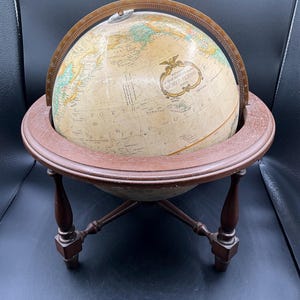 Może przedstawiać: Antyczny globus z kremową kulą, prezentujący cechy geograficzne w stonowanych odcieniach. Globus jest zamontowany na ciemnobrązowym drewnianym stojaku z czterema nogami. Na globusie widoczny jest napis "WORLD CLASS".