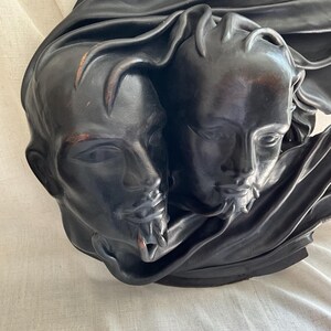 May include: A black and brown sculpture of two faces intertwined, with a flowing fabric draped over them. The sculpture is made of a smooth, textured material.