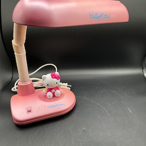 Puede incluir: Lámpara de escritorio rosa con una figura de Hello Kitty. La lámpara tiene un cuello flexible y una pantalla redondeada. La base y la pantalla tienen las palabras "Hello Kitty" en escritura azul. La figura es blanca con un lazo rosa.